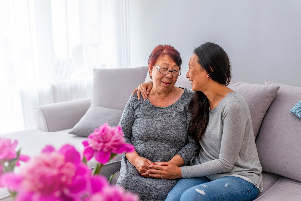 Close up family portrait of an adult Asian daughter with her mature mother leaning on each other and being affectionate while relaxing on a sofa at home.