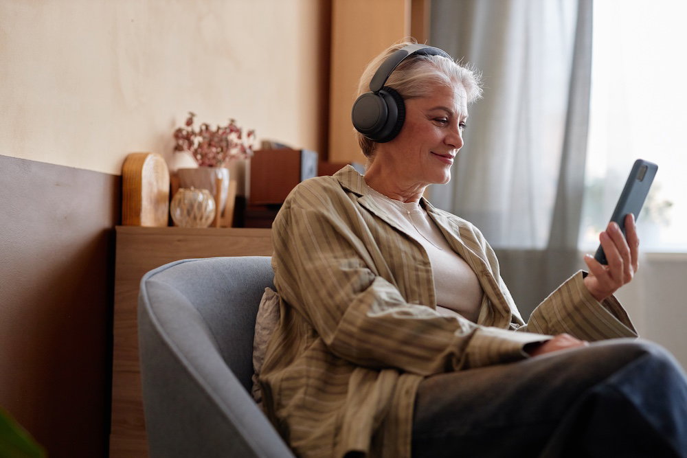Side view portrait of smiling grey haired senior woman listening to music and holding smartphone while sitting in armchair and enjoying relaxing day at home