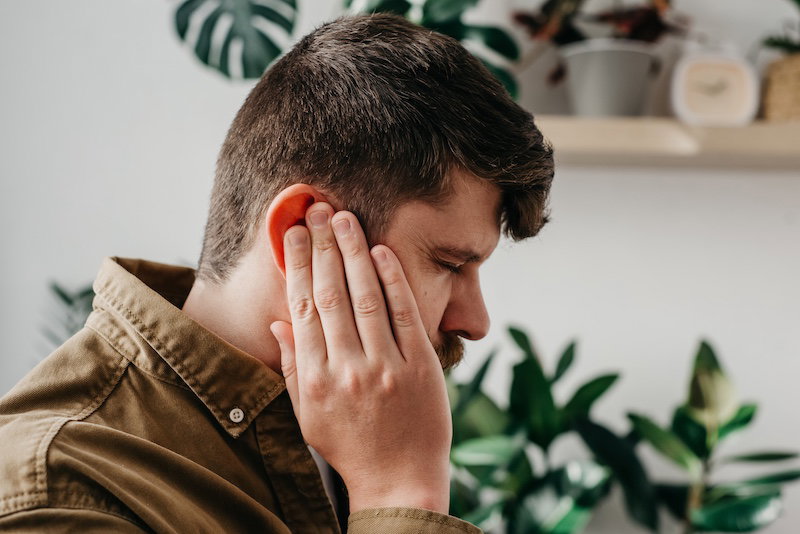 Close up of sick young caucasian male feeling ear pain, health care, neurological infection, itchiness otitis. Upset bearded man 30s old years having ear pain touching painful head sitting on couch